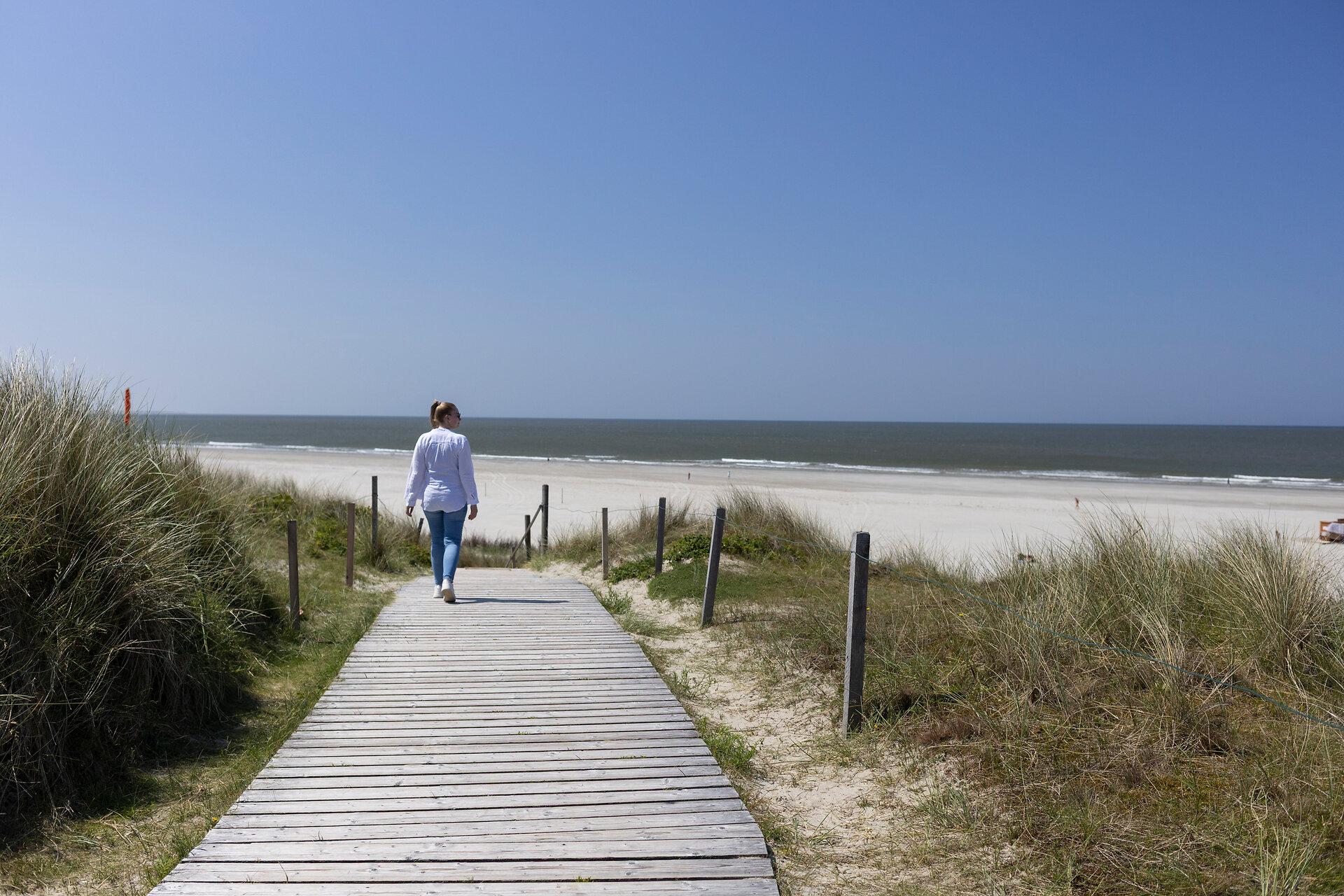 Frau spaziert entlang eines Holzbohlenwegs zum Strand auf Spiekeroog bei blauem Himmel