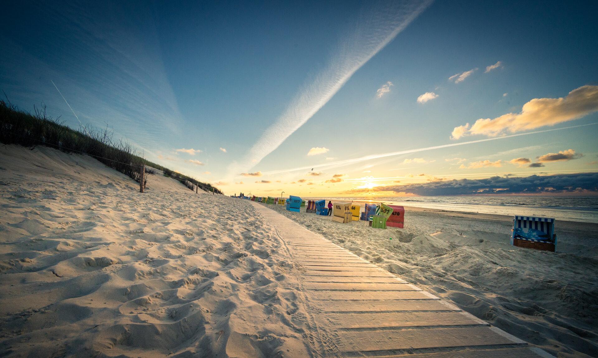 Ein Holzweg vorbei an Dünen und bunter Strandkörbe bei Sonnenuntergang am Strand von Langeoog
